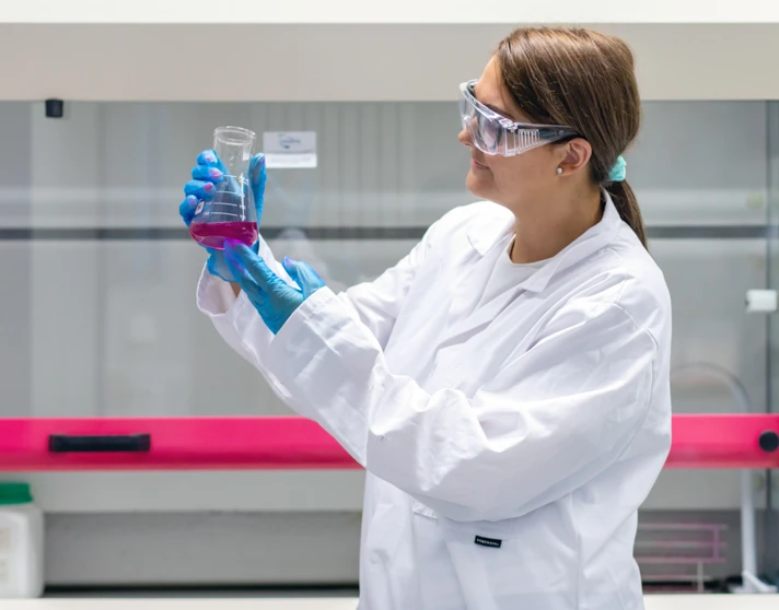 Student in a lab coat studying a beaker of pink liquid under bright lighting in a science lab. Student in a lab coat studying a beaker of pink liquid under bright lighting in a science lab.